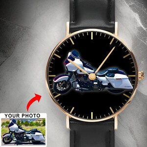 May include: A wristwatch with a black leather strap and a gold-tone case. The watch face features a black background with gold-tone hour markers and hands, displaying a graphic of a white touring motorcycle with blue accents and a fairing.