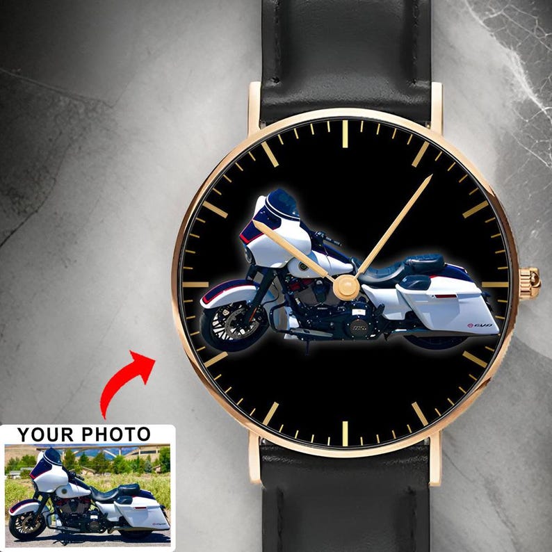 May include: A wristwatch with a black leather strap and a gold-tone case. The watch face features a black background with gold-tone hour markers and hands, displaying a graphic of a white touring motorcycle with blue accents and a fairing.