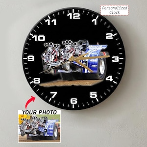May include: A round wall clock with a black face and white numbers. The center features a graphic of a blue tractor pulling sled with large engines. Gold-colored hour, minute, and second hands are centered. Text includes Personalized Clock.