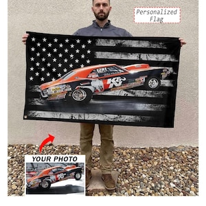 May include: A black flag with a distressed American flag design featuring a race car. The car is orange and black with racing decals. The flag has the text "Personalized Flag" in the upper right corner and "YOUR PHOTO" at the bottom.