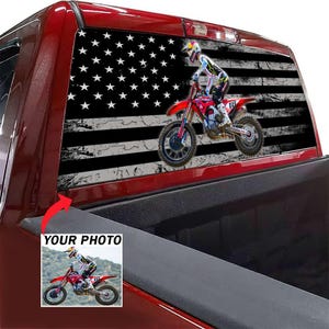 Peut inclure: Un autocollant pour lunette arrière de camion présentant un drapeau américain noir et blanc vieilli en arrière-plan avec un graphisme de pilote de motocross sur une moto rouge. Un petit encart montre la photo originale avec le texte YOUR PHOTO.