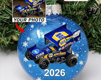May include: A round blue glitter Christmas bauble featuring a printed image of a blue and yellow NAPA Auto Parts sprint car with the number 49. The bauble displays the year 2026 in white text and includes a metal cap with a hanging loop.