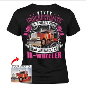 Puede incluir: Camiseta negra con un diseño de camión de 18 ruedas rojo. La camiseta muestra el texto "NEVER UNDERESTIMATE THE POWER OF A WOMAN WHO CAN HANDLE AN 18-WHEELER" en rosa y blanco. Una imagen más pequeña del camión está en la esquina.