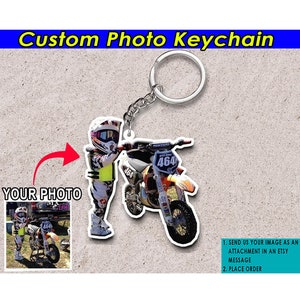May include: A custom photo keychain featuring a child in a motocross racing suit holding a dirt bike. The keychain is made of clear acrylic and has a silver metal key ring.