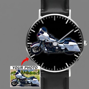 May include: A round analog wristwatch with a black leather strap and a silver-toned case. The watch face features a black background with a white motorcycle graphic and silver hands. A small inset shows a photo of the same white motorcycle.