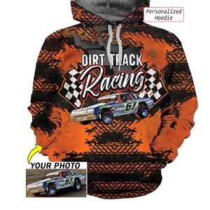 May include: A hooded sweatshirt featuring a vibrant orange and black dirt track racing graphic with tire tread patterns, checkered flags, and the text "DIRT TRACK Racing". The design includes a central race car numbered 67 and a custom photo insert.