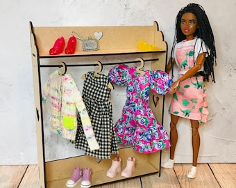 May include: A wooden doll clothes rack with four hangers holding a pink floral dress, a black and white gingham dress, a white and pink sweater, and a pink and white floral dress. A doll with brown hair and a pink and white dress is standing next to the rack.