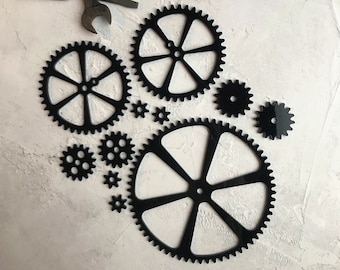 以下が含まれることがあります： 白い背景に、さまざまなサイズの黒い金属製の歯車、6本のスポークを持つ大きな歯車などが配置されています。