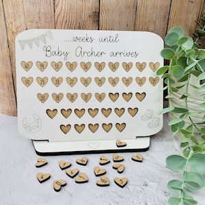May include: A white wooden pregnancy countdown board with the text "...weeks until Baby Archer arrives." It features heart-shaped cutouts for tracking weeks, with wooden heart-shaped number tokens. Includes baby-themed illustrations.