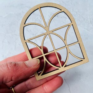 Op de afbeelding: Een houten uitsnijding van een gebogen raam met een geometrisch ontwerp. Het raam is gemaakt van dun hout en heeft een lichtbruine kleur.