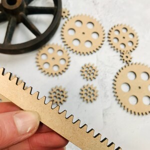 以下が含まれることがあります： さまざまなサイズの木製ギアのセット。長い薄い歯付きギアも含まれています。ギアは茶色で、黒い縁取りがされています。ギアは白い表面にあります。