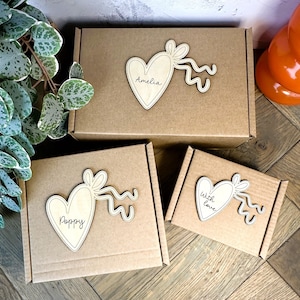 May include: Three brown cardboard boxes of varying sizes, each adorned with a wooden heart-shaped embellishment. The hearts have a ribbon design and personalized names or phrases: "Amelia", "Poppy", and "With Love".