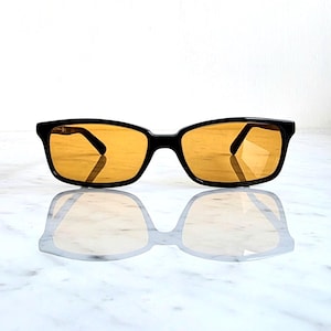 Puede incluir: Gafas de sol negras con lentes ámbar. Las monturas rectangulares tienen un acabado brillante y las lentes son de un color dorado cálido. Las gafas de sol se reflejan en una superficie blanca.