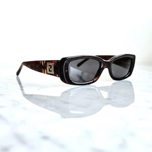 Può includere: Occhiali da sole rettangolari con montatura a fantasia tartarugata e lenti scure. Gli occhiali da sole hanno un'emblema dorato sulle aste. Gli occhiali si riflettono su una superficie bianca.