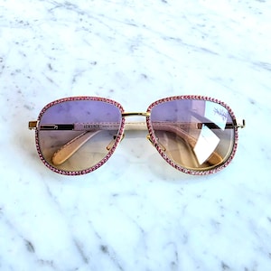 May include: Gold-framed sunglasses with pink and purple gradient lenses. The frames are adorned with pink gemstones around the lenses. The brand name "VERSACE" is visible on the left arm. Made in Italy.