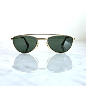 May include: Gold-framed sunglasses with dark green lenses. The sunglasses have a double bridge and a classic aviator style. The lenses are oval-shaped and reflect the light. The sunglasses are displayed on a white surface.