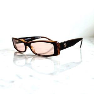 May include: Rectangular eyeglasses with a brown and black frame and pink-tinted lenses. The glasses have a thick frame with a small decorative emblem on the temple. The lenses are a light pink color.