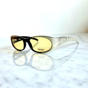 May include: Versace sunglasses with a black frame and yellow lenses. The temples feature a snakeskin pattern in white and gray. The brand name "VERSACE" is printed on the lens.