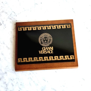 May include: A rectangular wooden tray with a black center featuring the gold Gianni Versace logo and text. The tray is framed with a gold Greek key pattern.