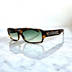 Könnte beinhalten: Schildpatt-Sonnenbrille mit grünen Farbverlaufsgläsern. Der Markenname "CHANEL" ist mit funkelnden Verzierungen auf den Bügeln geschrieben. Die Sonnenbrille spiegelt sich auf einer Marmoroberfläche.