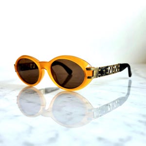 Op de afbeelding: Ovale zonnebril met een doorschijnend oranje montuur en donkere glazen. De veren hebben een goudkleurig kettingontwerp met een logo. De zonnebril wordt weerspiegeld op een wit oppervlak.