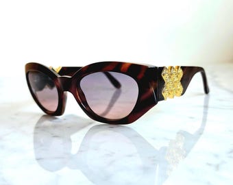May include: Brown tortoiseshell sunglasses with a cat-eye shape and gold-colored embellishments on the temples. The lenses have a gradient tint, transitioning from a darker shade at the top to a lighter hue at the bottom. The sunglasses are set against a white background.