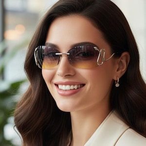 May include: A woman wearing rimless sunglasses with gradient lenses and gold-colored frames. She is wearing a cream-colored blazer and a white blouse. The background includes a window, a plant, and a light fixture.