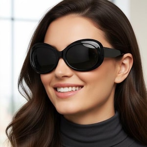 May include: Black oval sunglasses with thick frames. The sunglasses are worn by a person with long, dark hair and a dark gray turtleneck. The person is smiling.