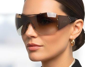 May include: Brown-tinted sunglasses with a shield design. The sunglasses have a dark brown frame and a gradient lens. The model is wearing a black blazer and a white blouse. Gold hoop earrings are visible.