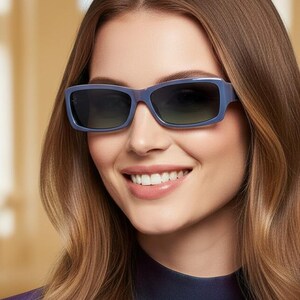 May include: A person wearing blue rectangular sunglasses with dark lenses. The sunglasses have a thick frame and are set against a blurred background. The person is smiling and wearing a blue and purple top.