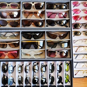 May include: A collection of various styles of sunglasses, including aviator, wayfarer, and round frames, displayed in a wooden case. The sunglasses are arranged in rows, with different colours and designs.