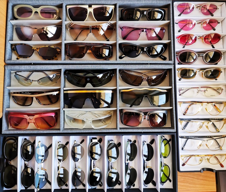 May include: A collection of various styles of sunglasses, including aviator, wayfarer, and round frames, displayed in a wooden case. The sunglasses are arranged in rows, with different colours and designs.