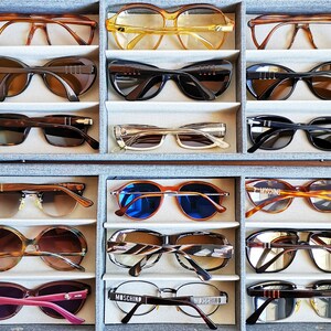 Puede incluir: Una colecci&oacute;n de gafas de sol vintage en varios estilos y colores, incluyendo monturas marrones, negras, amarillas y rosas. Las gafas de sol est&aacute;n dispuestas en un organizador gris, mostrando una variedad de formas y dise&ntilde;os, desde cl&aacute;sicos hasta audaces.
