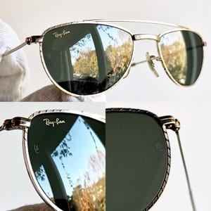 May include: Gold-framed Ray-Ban sunglasses with green lenses. The lenses reflect a landscape scene. The frames have a double bridge and decorative detailing. The Ray-Ban logo is visible on the top left lens.