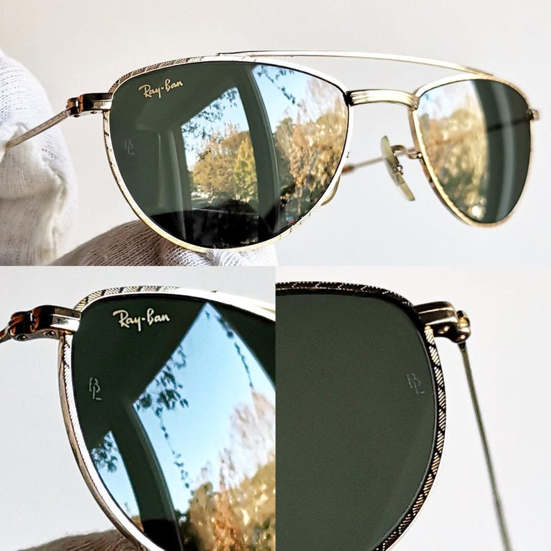 May include: Gold-framed Ray-Ban sunglasses with green lenses. The lenses reflect a landscape scene. The frames have a double bridge and decorative detailing. The Ray-Ban logo is visible on the top left lens.
