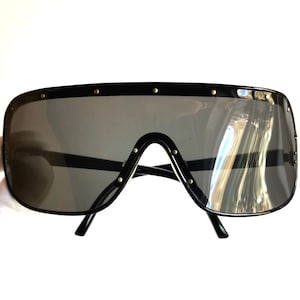 May include: Black oversized sunglasses with a shield-style design. The sunglasses have a dark gray lens and are accented with small gold-colored studs along the top and bridge. The frame is a glossy black.