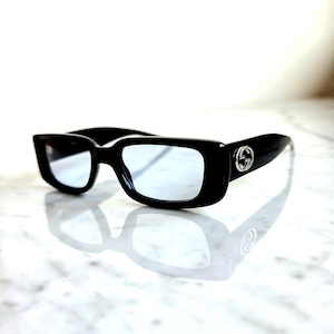May include: Black rectangular sunglasses with light blue lenses. The sunglasses have a thick frame and a silver logo on the temples. The glasses are reflected on a white surface.