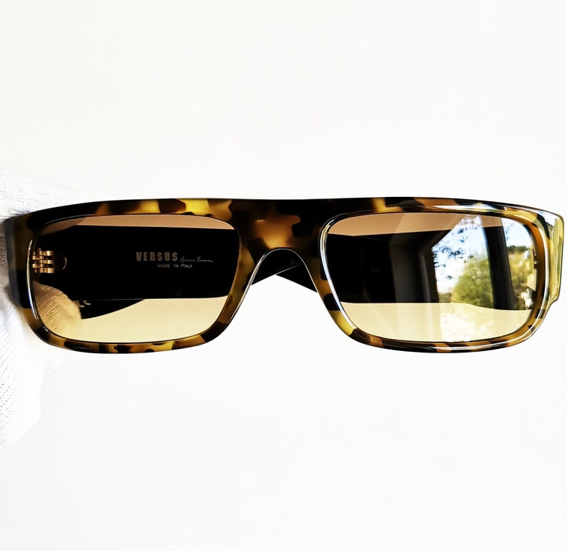 May include: Tortoiseshell sunglasses with brown lenses. The sunglasses have a square frame and the brand name 'VERSUS' is printed on the left lens.