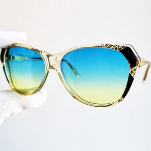 May include: Clear-framed sunglasses with gradient blue and yellow lenses. The sunglasses have a cat-eye shape with black and gold accents on the outer corners and along the top frame. The lenses transition from yellow to blue.