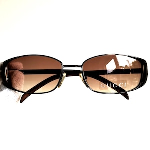 May include: Brown gradient lens sunglasses with a dark brown metal frame. The sunglasses have a rectangular shape and the brand name "GUCCI" is visible on the lens.