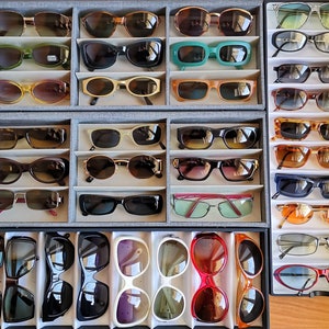May include: A display of various sunglasses in different colours and styles, arranged in rows on a grey background. The sunglasses are organised by colour and style, with some featuring mirrored lenses and others with clear lenses.