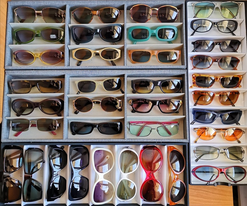 May include: A display of various sunglasses in different colours and styles, arranged in rows on a grey background. The sunglasses are organised by colour and style, with some featuring mirrored lenses and others with clear lenses.