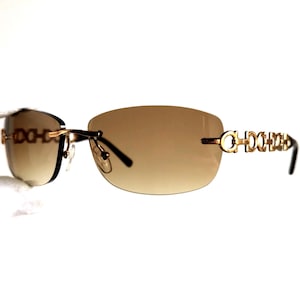 May include: Rimless sunglasses with brown gradient lenses and gold-tone metal accents. The temples feature a repeating chain design. The sunglasses have a classic, elegant style.