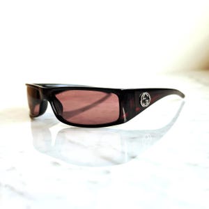 May include: Dark brown rectangular sunglasses with tinted lenses. The sunglasses have a silver emblem on the temples. The lenses are a reddish-brown color. The sunglasses are on a white surface.