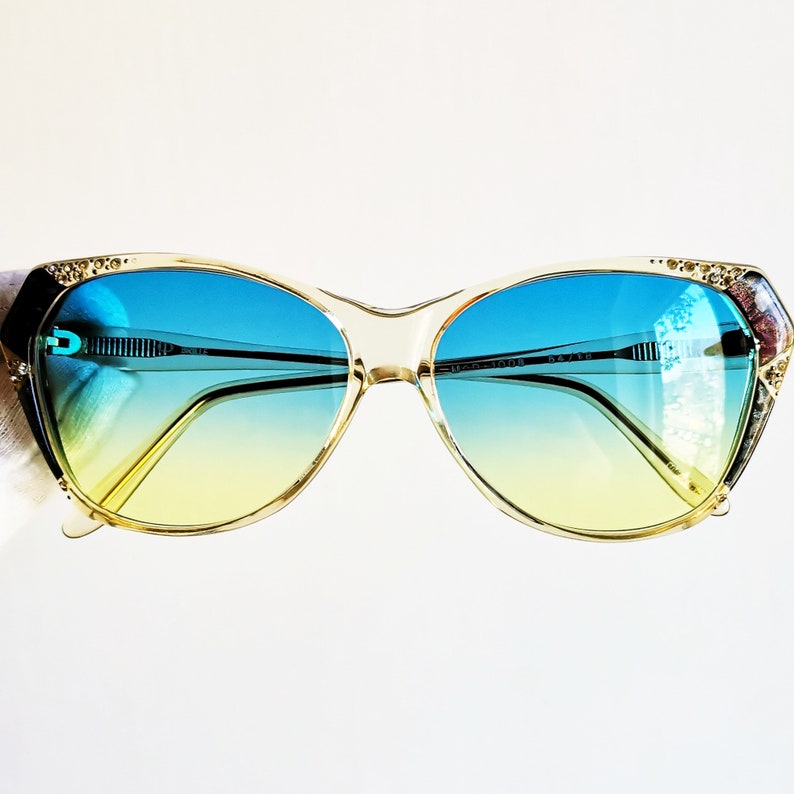 May include: Vintage cat-eye sunglasses with a gradient blue to yellow lens. The frames are a light beige color with gold accents and decorative rhinestones on the outer corners. The sunglasses have a classic, retro style.