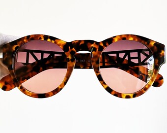 May include: Tortoiseshell sunglasses with a black frame and pink lenses. The frame has a geometric design.
