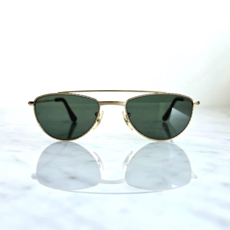 May include: Gold-framed sunglasses with dark green lenses. The sunglasses have a double bridge and a classic aviator style. The lenses are oval-shaped and reflect the light. The sunglasses are displayed on a white surface.