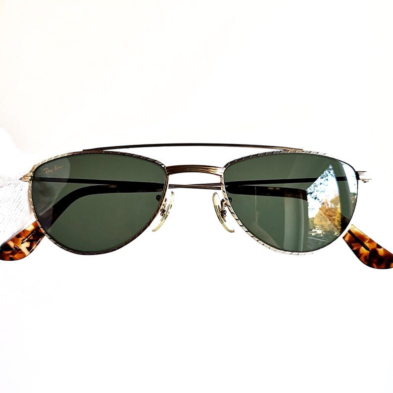 May include: Gold-framed sunglasses with dark green lenses and a double bridge. The temples feature a tortoiseshell pattern. The Ray-Ban logo is visible on the upper left lens. These sunglasses offer a classic, stylish look.