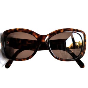 Puede incluir: Gafas de sol marrones con estampado de carey y lentes marrones. Las gafas tienen forma rectangular redondeada y montura gruesa. Un accesorio de moda.