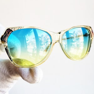 May include: Clear-framed sunglasses with blue and yellow gradient lenses. The frames have decorative detailing on the upper corners. The lenses reflect a bright outdoor scene. A stylish accessory for eye protection.